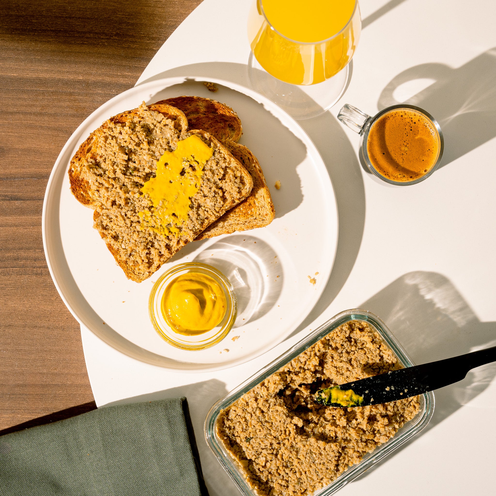 Petit déjeuner composé de tranches de pain garnies de cretons végétaux Grounded et de moutarde accompagné d’un café espresso et d’un verre de jus d’orange. Protéine végétale de pois servie dans un plat en verre avec spatule.