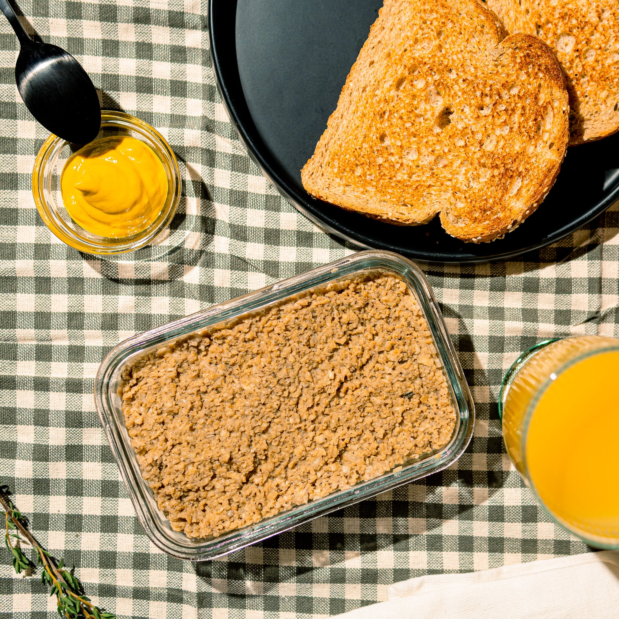 Scène de petit déjeuner avec une tartinade végétale de type cretons signée Grounded servie dans un plat en verre. À côté on retrouve des tranches de pain grillé de la moutarde du jus d’orange et une nappe à carreaux style champêtre.
