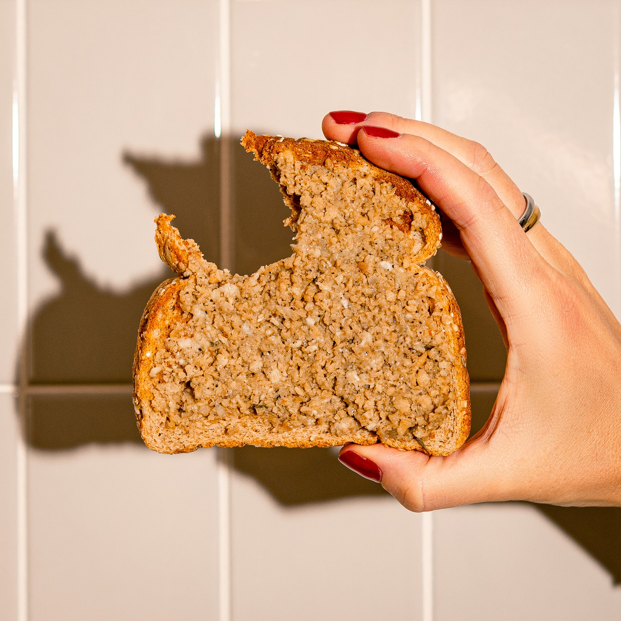 Tranche de pain garnie de cretons végétaux Grounded tenue en main avec une bouchée croquée. Texture onctueuse à base de protéine végétale de pois présentée devant un mur de céramique blanche.