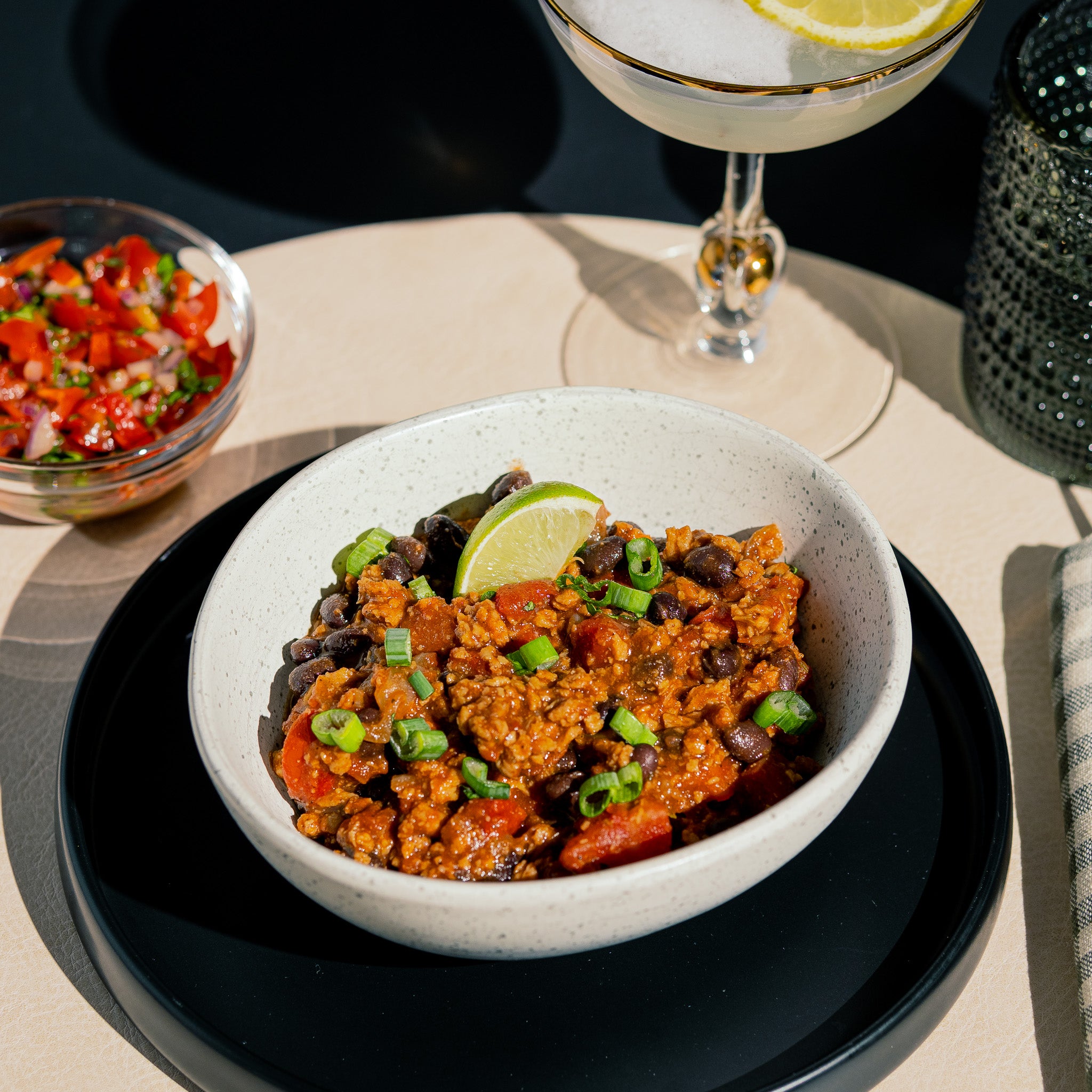 Bol de chili végétalien cuisiné avec la protéine Texmex de Grounded. Garnie de coriandre d’oignons verts de haricots noirs et de lime la recette est servie avec un cocktail et une salsa fraîche.