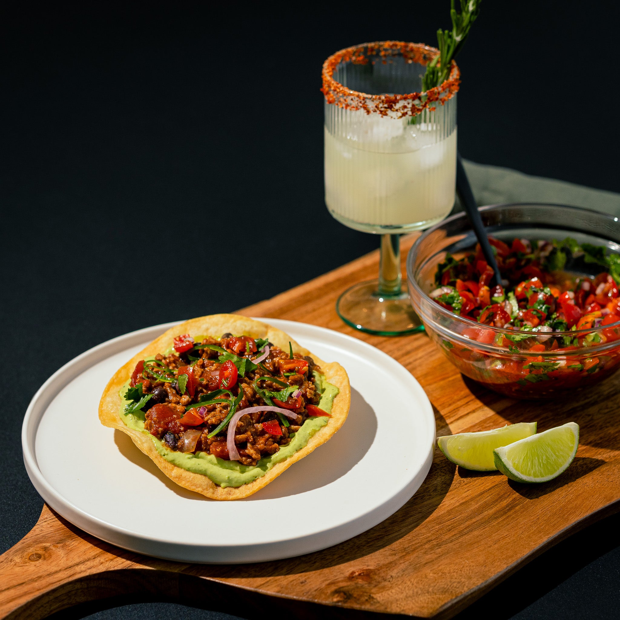 Tostada garnie de guacamole et de protéine végétale Texmex Grounded avec salsa fraîche oignons rouges coriandre et lime. Servie avec un cocktail givré sur une planche en bois pour une présentation estivale.